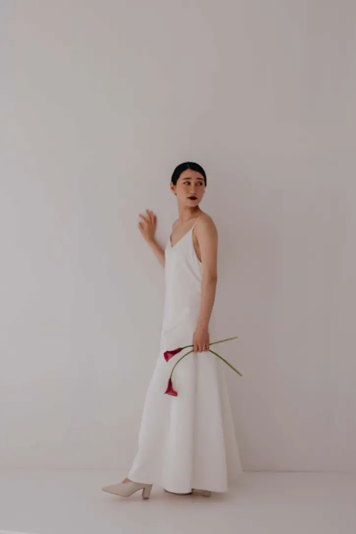 白いキャミソールドレスを着た女性が赤いカラーの花を持って立つ写真