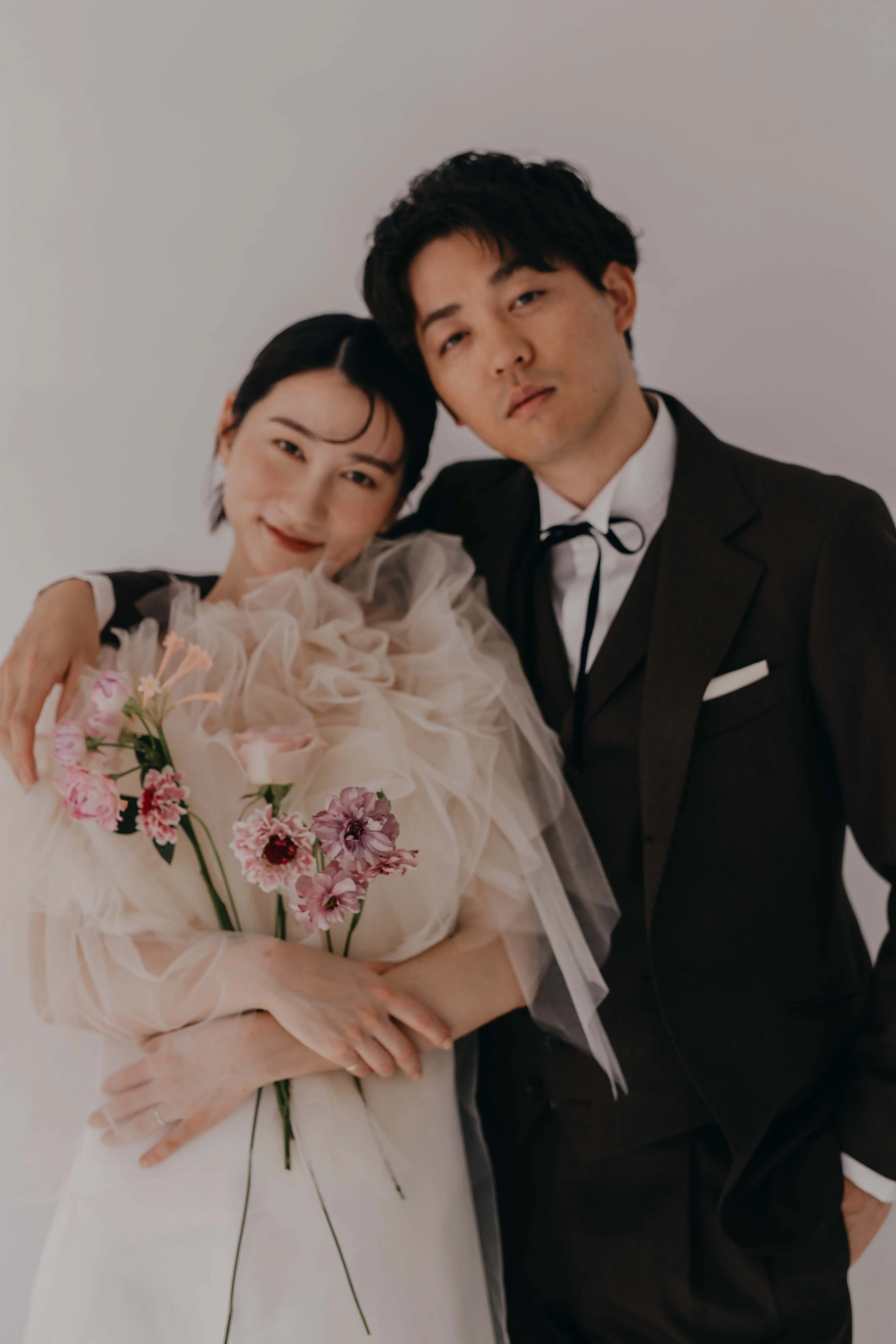 チュール素材の白いドレスを着てピンクの花を抱えた女性に、ダークスーツにボウタイの男性が肩に手をまわして寄り添うブライダルフォト
