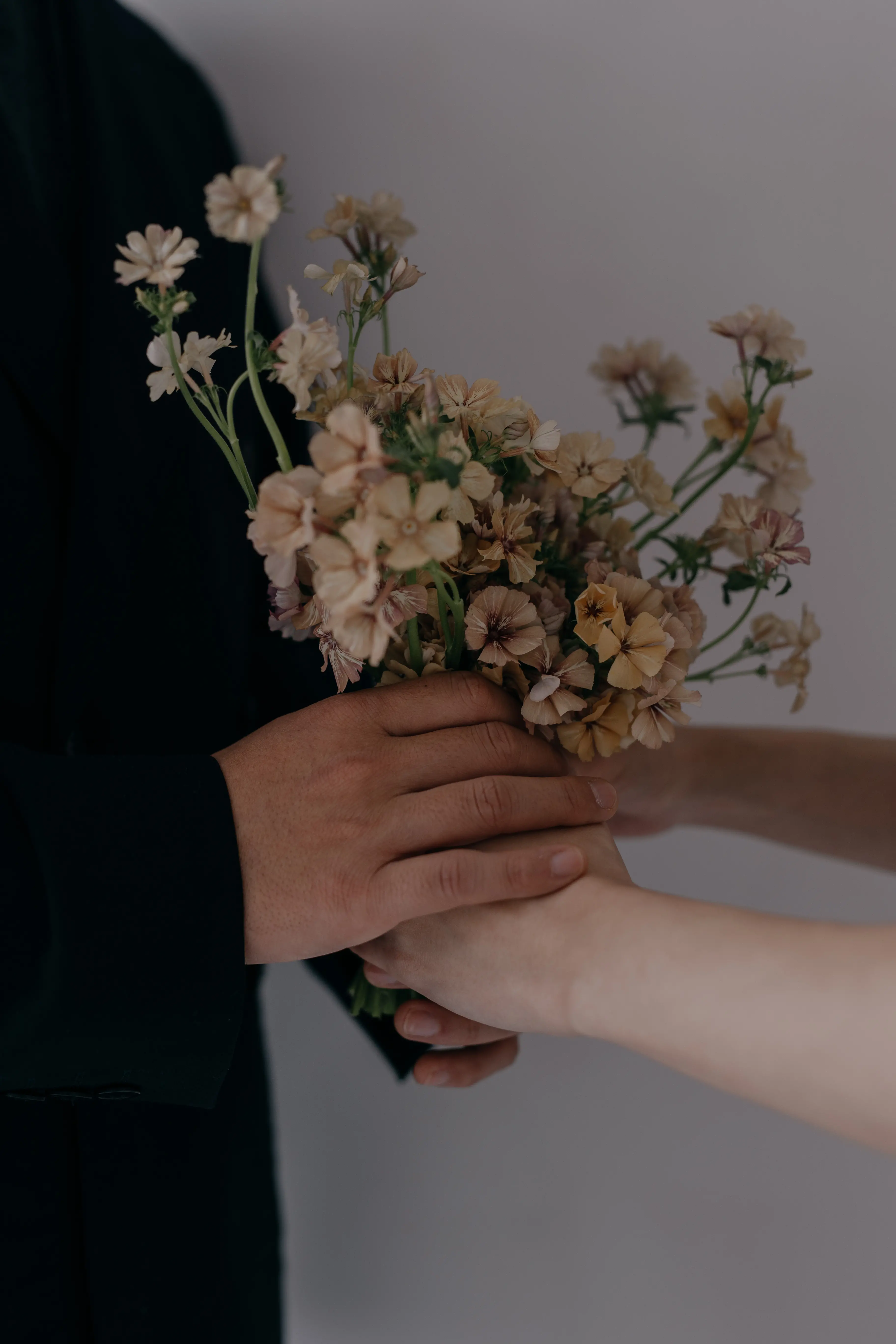 ダークスーツの男性と白い衣装の女性が共に小さなベージュの花束を手で包むように持つ、暗い背景のクローズアップ