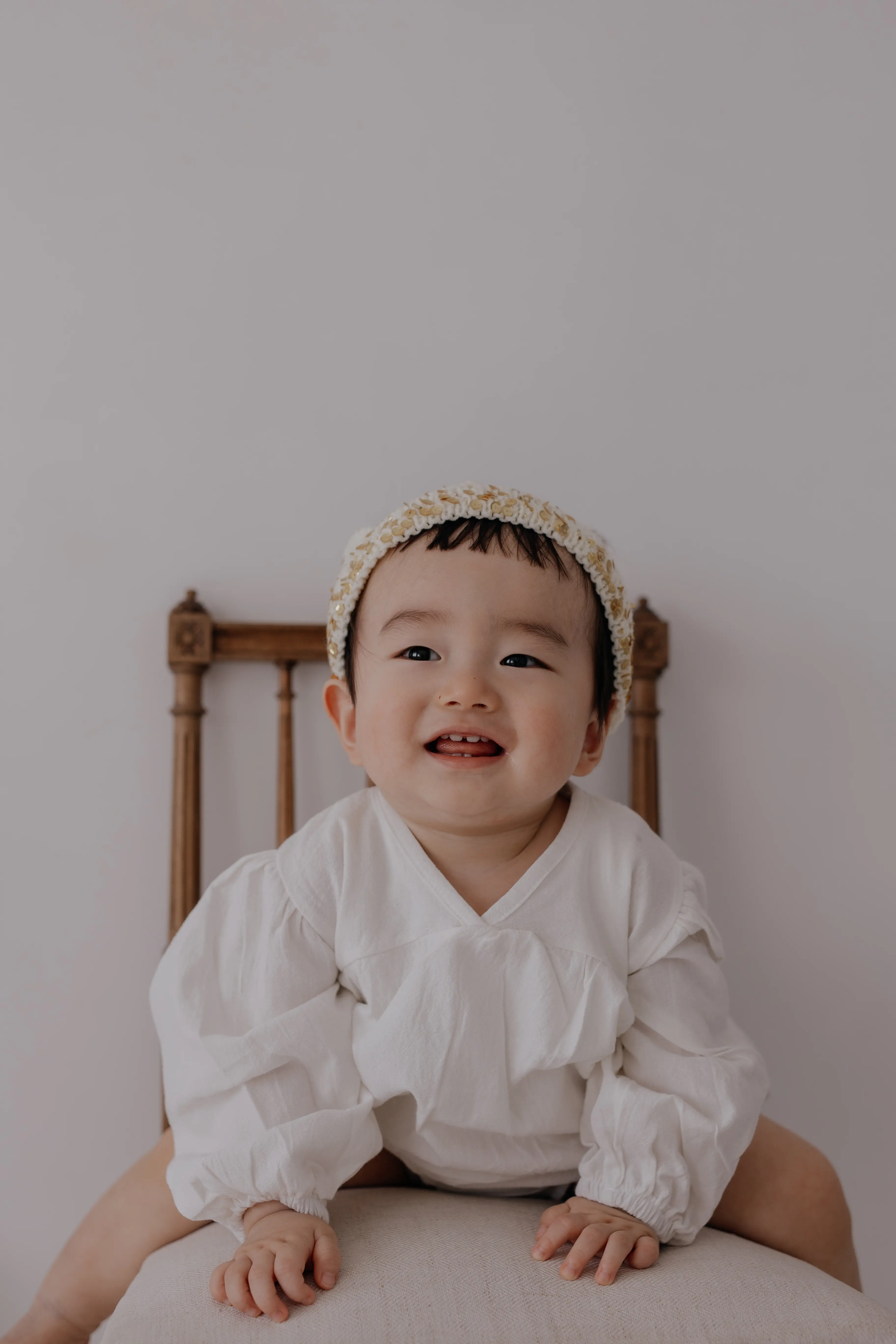 白い服とフラワークラウンをつけた赤ちゃんが椅子に座り、笑顔を向けているポートレート