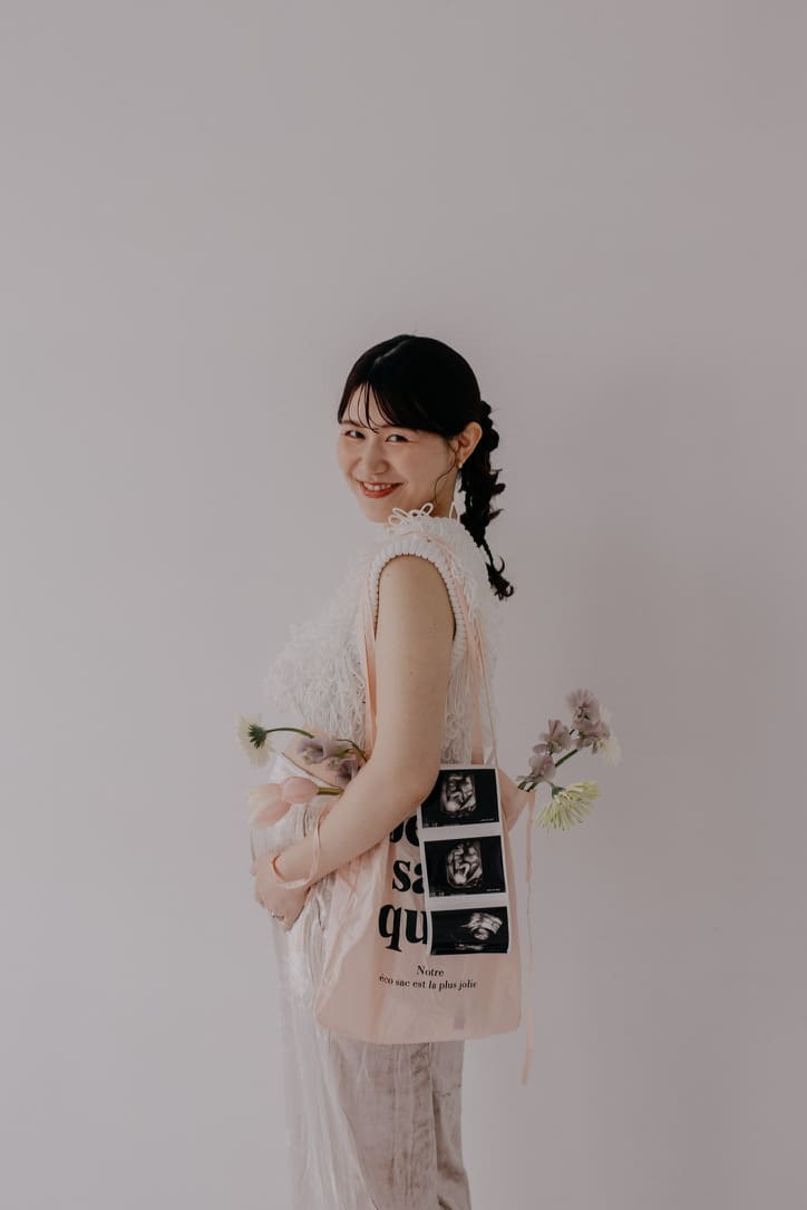 振り返って微笑む妊婦さんがエコー写真のプリントされたトートバッグと花を持つマタニティフォト